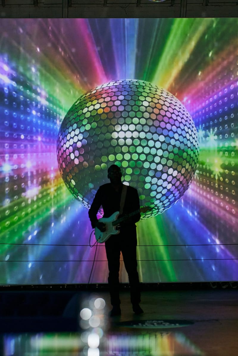 A guitarist silhouetted against a huge rainbow disco ball video wall