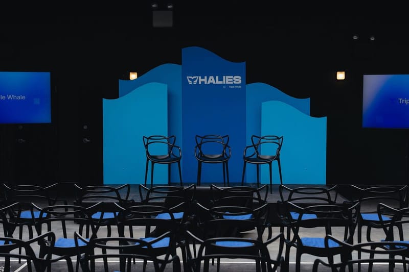 Three black chairs on a small stage before blue wave-shaped panels with the Whalies logo, facing rows of seats